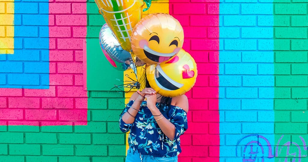 "A woman concealing her face behind balloons with emoji faces, standing against a vibrant, colorful wall