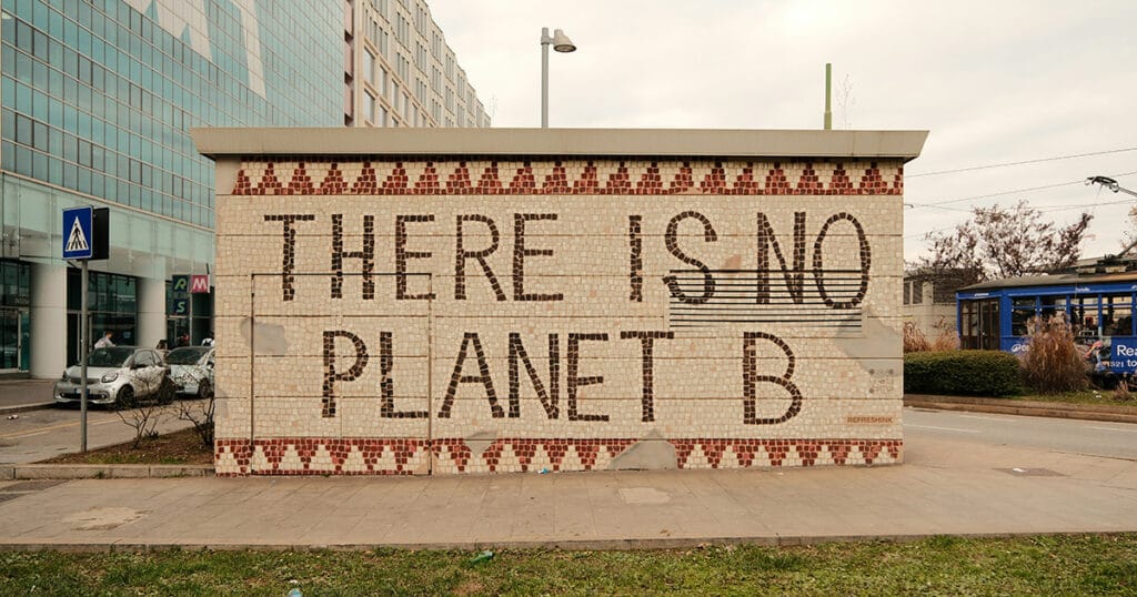 A large tiled wall in an urban street setting with the message “THERE IS NO PLANET B” written in bold capital letters. The wall has decorative red and cream patterned tiles along the top and bottom edges. In the background, modern glass buildings, parked cars, a tram, and pedestrians are visible under a cloudy sky.