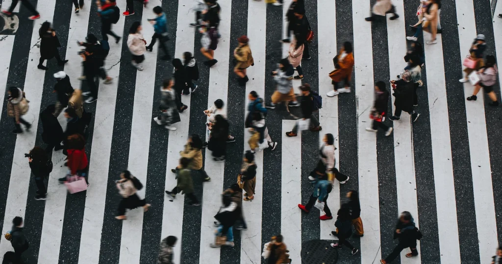 Aerial view of a busy city crosswalk with black-and-white stripes, showing dozens of pedestrians in colorful clothing moving in different directions with motion blur, creating a dynamic sense of urban energy.